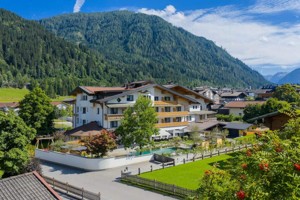Außenaufnahme der Rückseite des Hotels samt Garten, Kinderspielplatz und Naturbadeteich